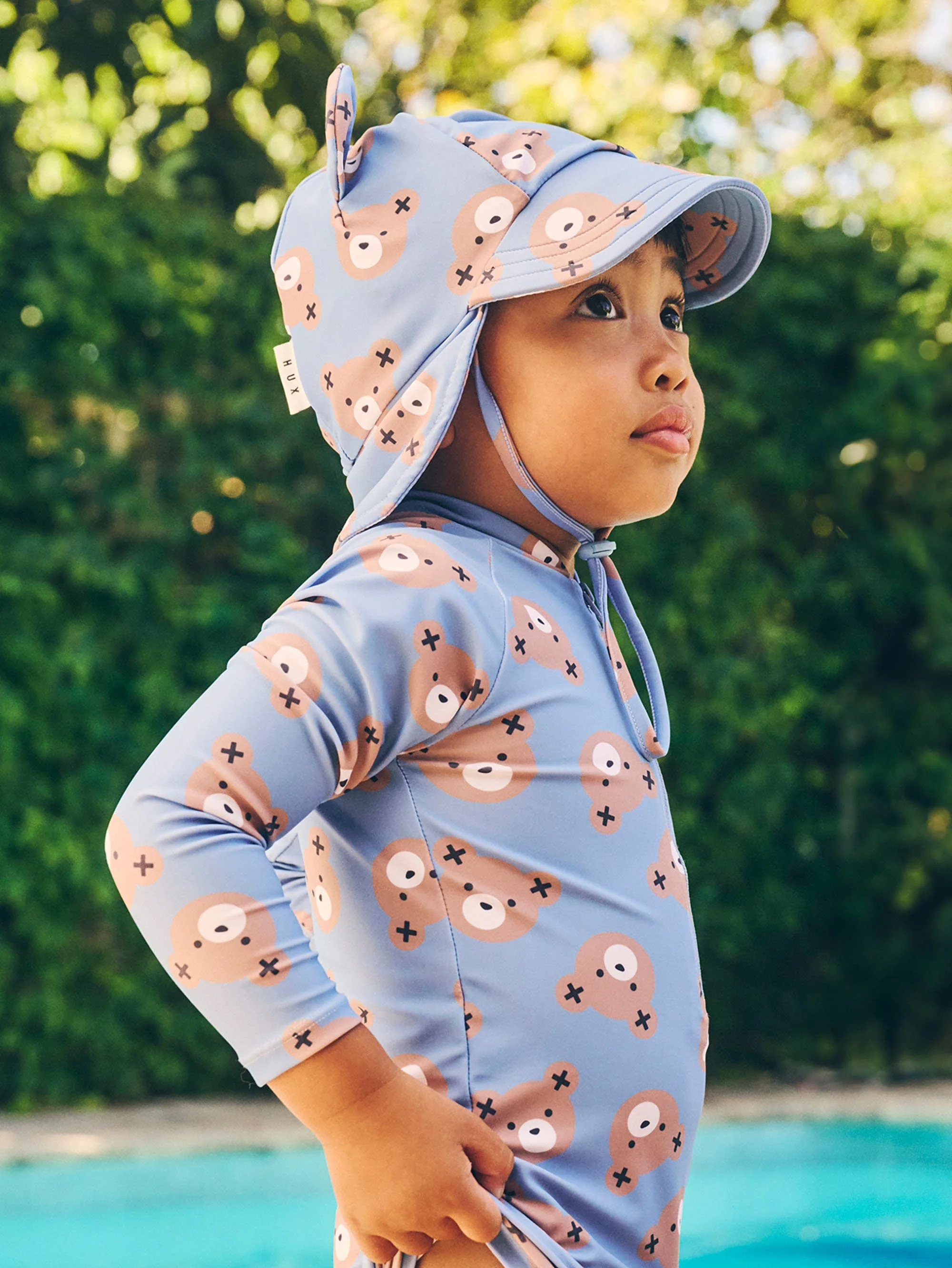 Child in bear-print swimwear and sun hat near a pool with green foliage backdrop. Outdoor setting, eye-level view, bright natural light.