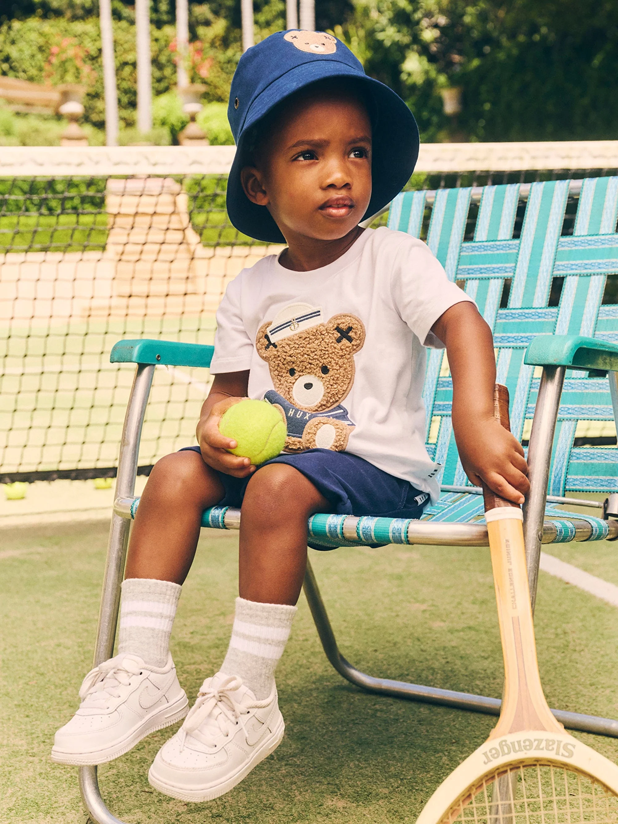 Young boy in bucket hat and teddy bear t-shirt sitting on a light blue tennis chair with a tennis ball and racquet in hand.