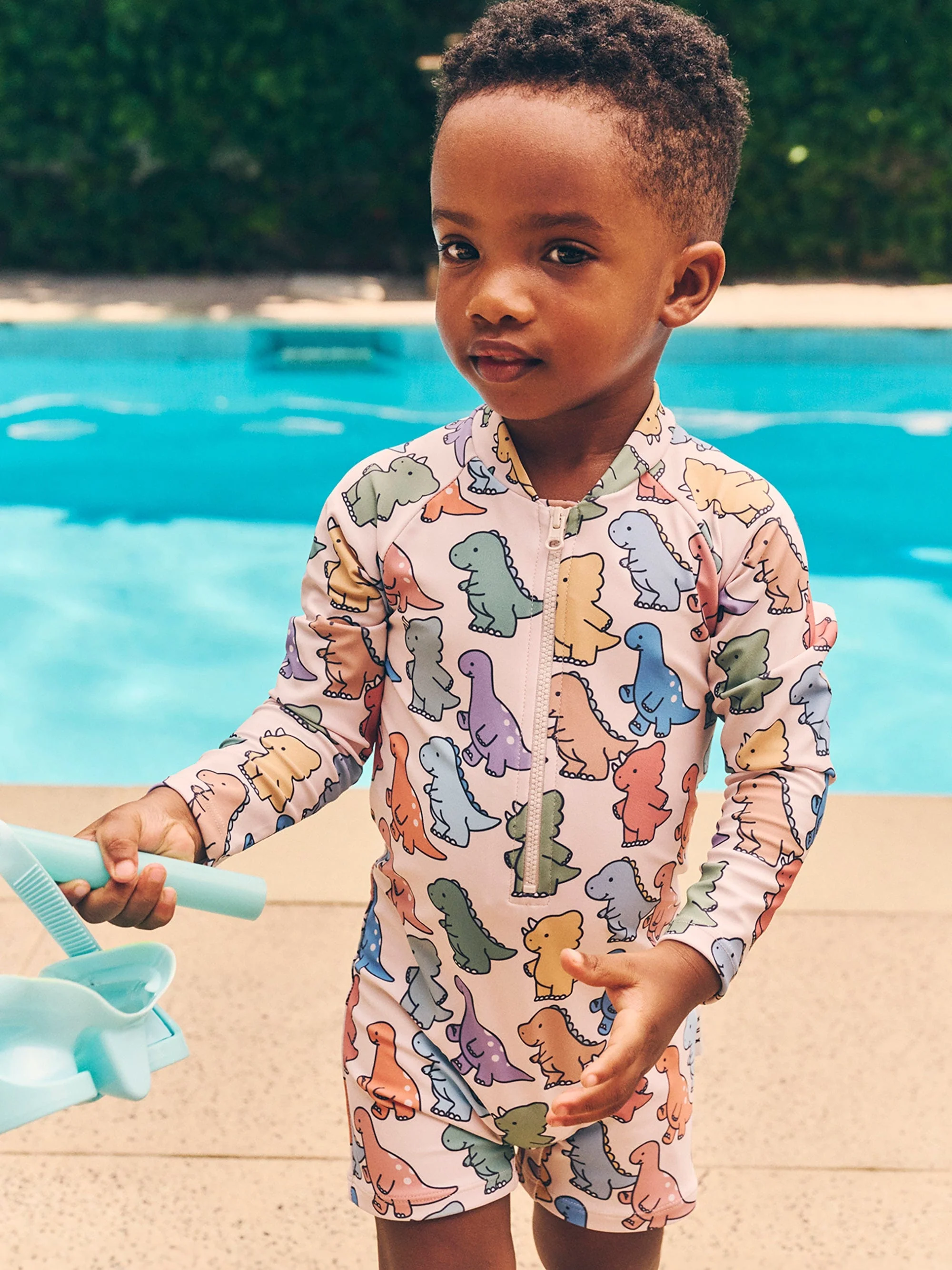 A young child stands near a pool wearing a dinosaur-print swimsuit and holding a blue toy shovel and rake. Outdoor summer scene.