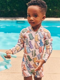 Young child in a dinosaur print rashguard swimsuit holding a toy by an outdoor swimming pool