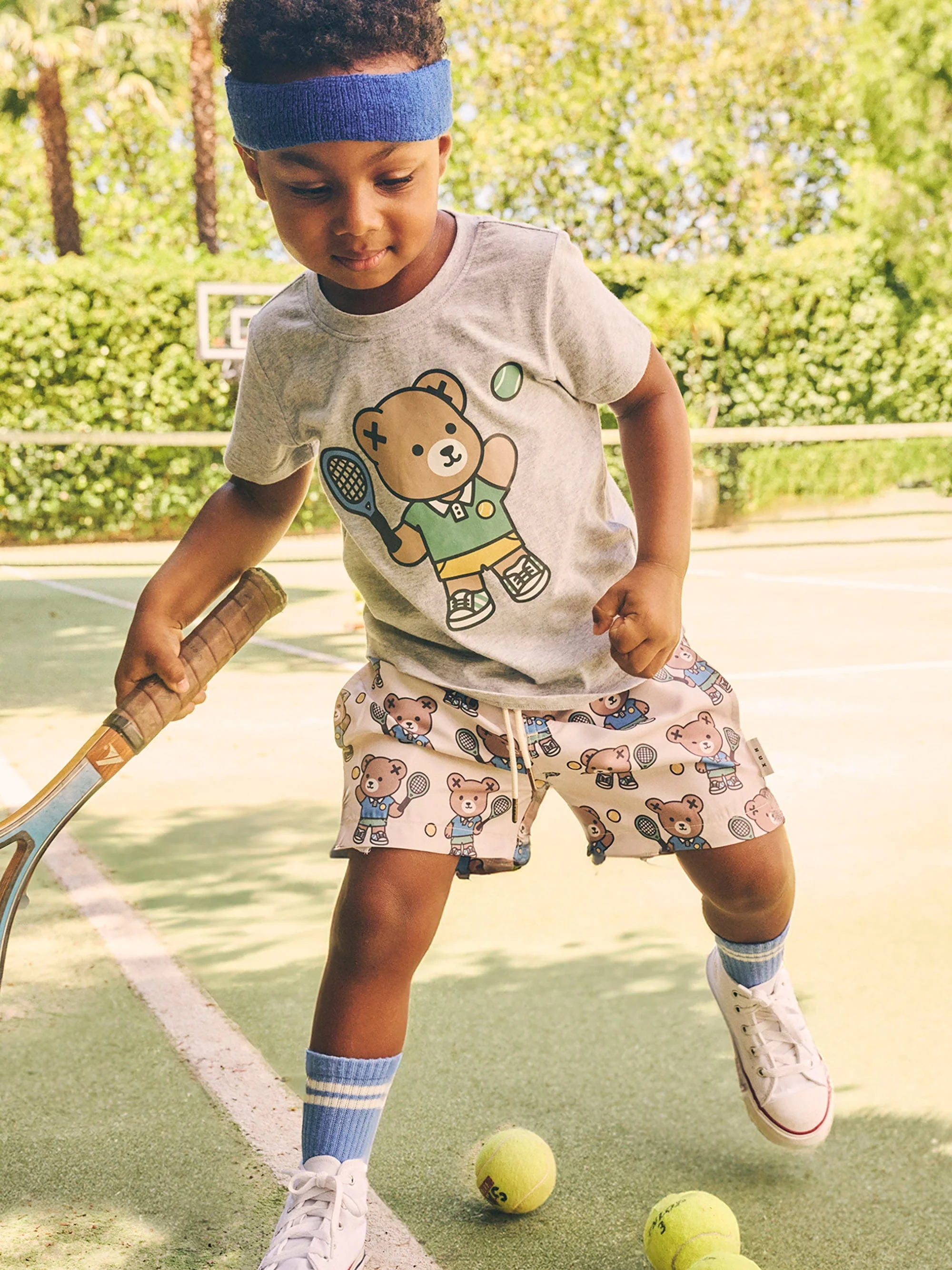 Young child playing tennis on a green tennis court, wearing a blue headband, t-shirt with teddy bear design, and tennis-themed shorts.