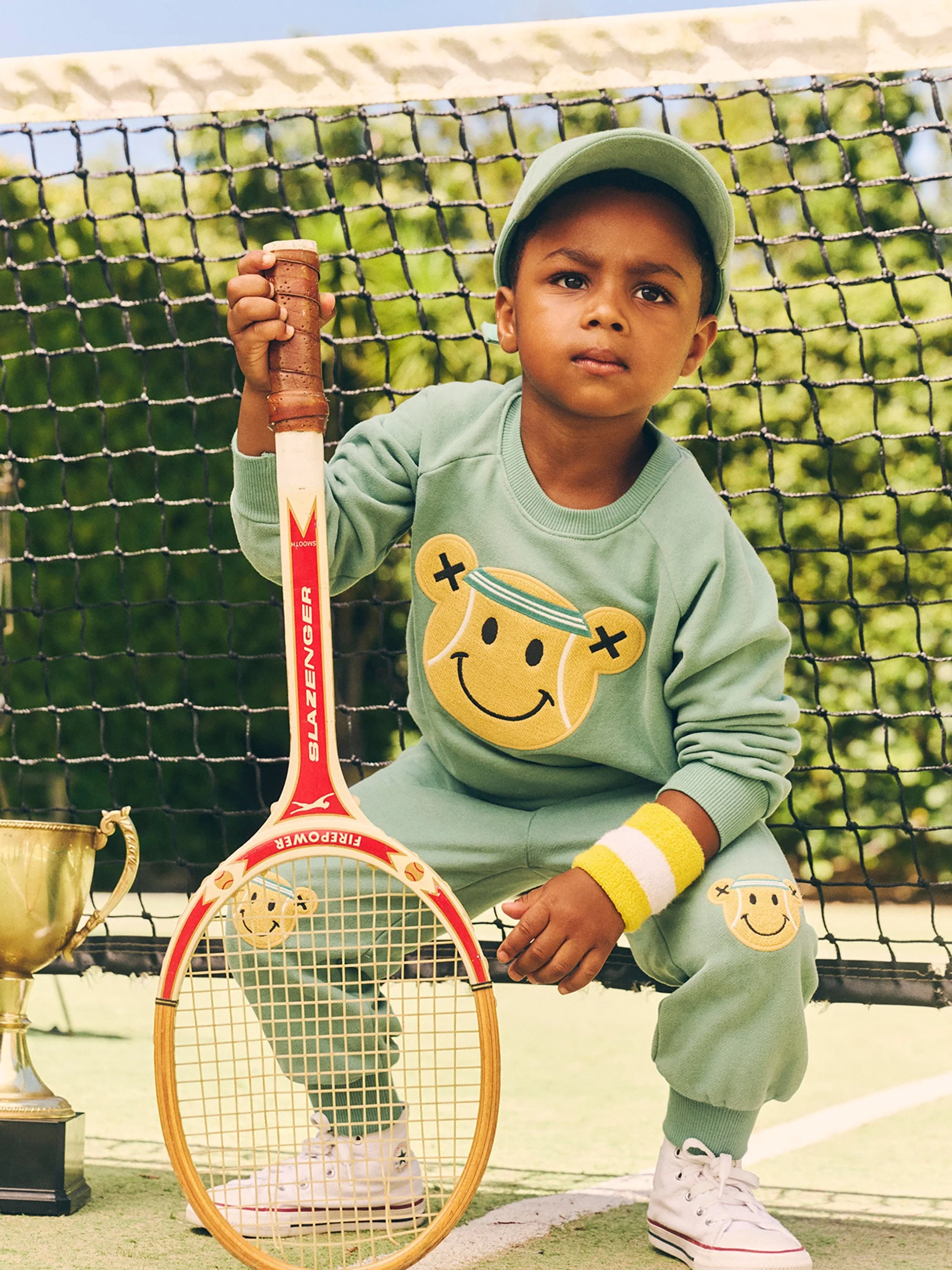A child in a green sweatsuit holds a tennis racket while posing on a tennis court next to a trophy, creating a charming, sporty scene.