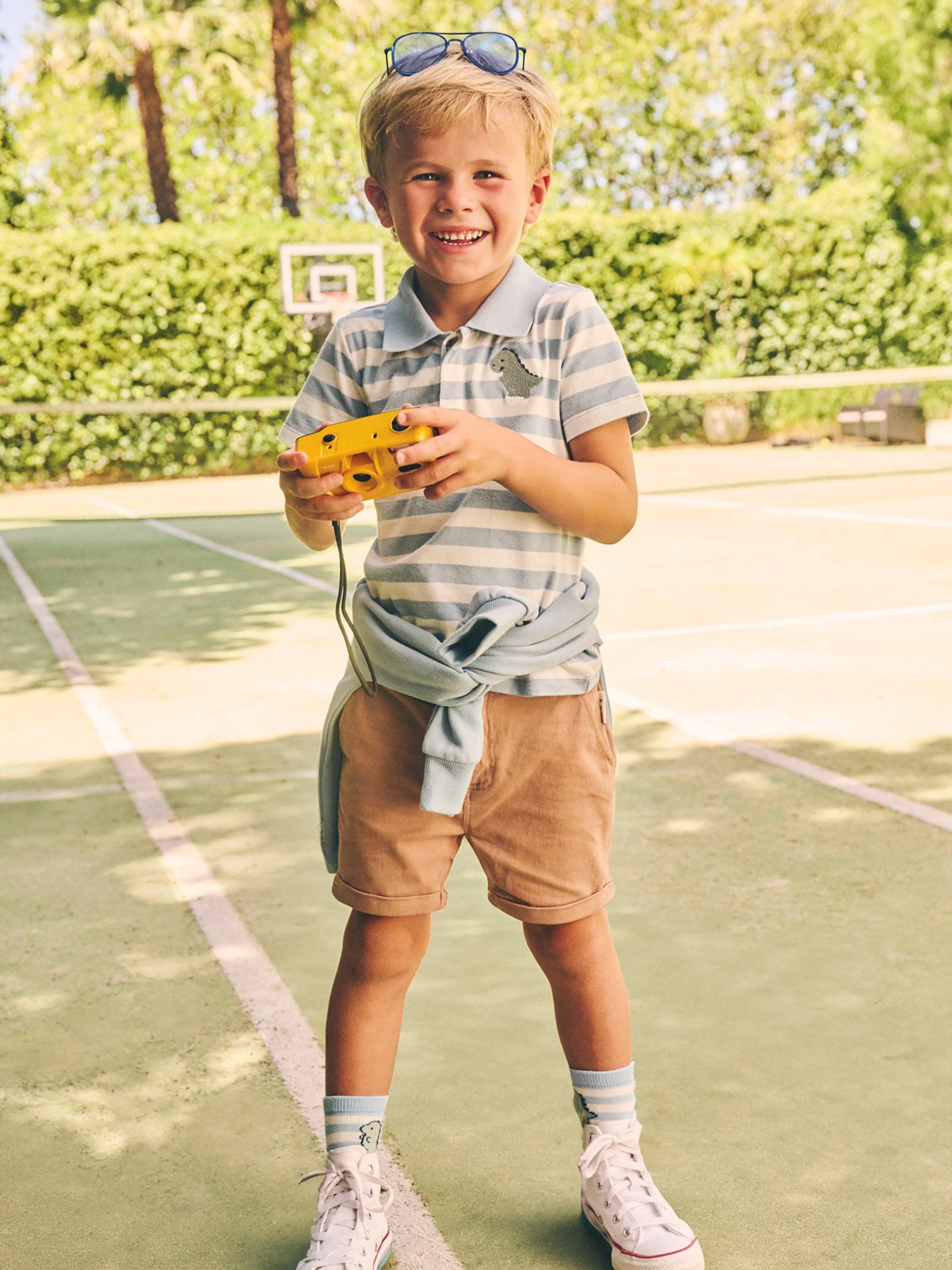 Young boy smiles on tennis court, holding yellow toy camera with sunglasses on his head, wearing striped shirt and shorts outdoors.