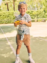 Young child holding a yellow toy camera on a tennis court, wearing a striped polo shirt and brown shorts.