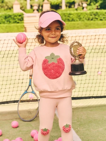 A young girl smiles holding a trophy and tennis ball on a tennis court, wearing a pink strawberry-themed outfit and visor.