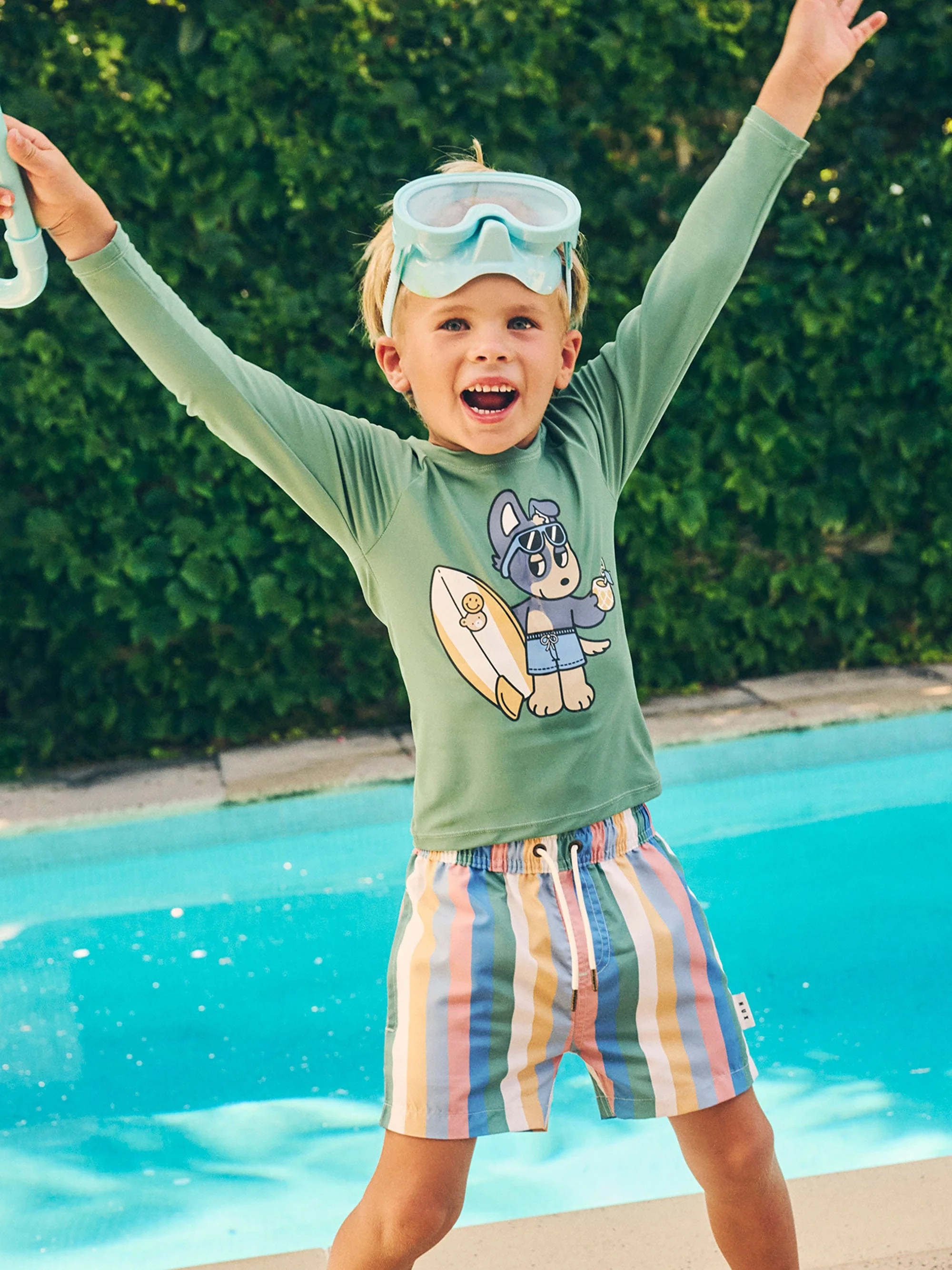 Young child wearing a green long-sleeve rash guard and striped swim shorts standing outdoors by a blue swimming pool with snorkel goggles.