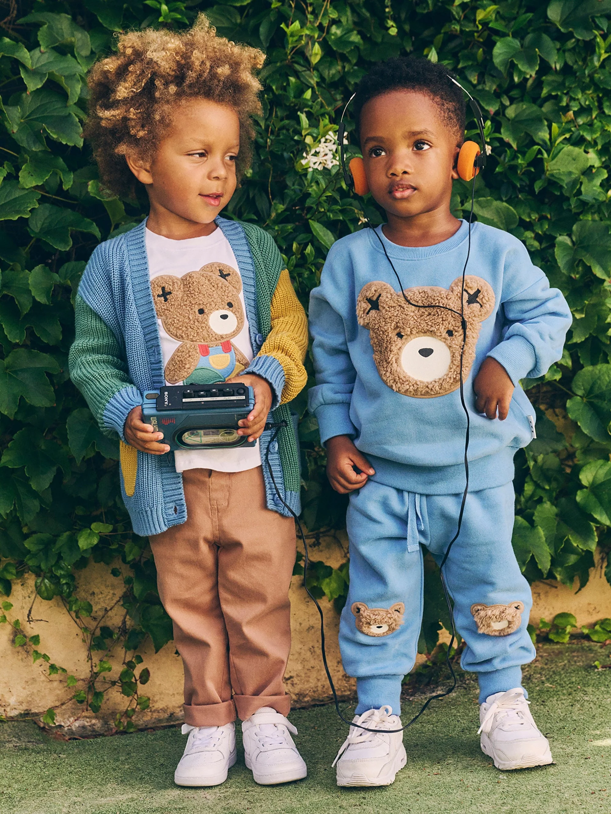 Two young children wearing matching teddy bear themed outfits stand in front of green foliage, one holding a cassette player.