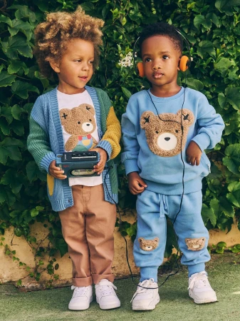 Two young children wearing matching teddy bear themed outfits stand in front of green foliage, one holding a cassette player.