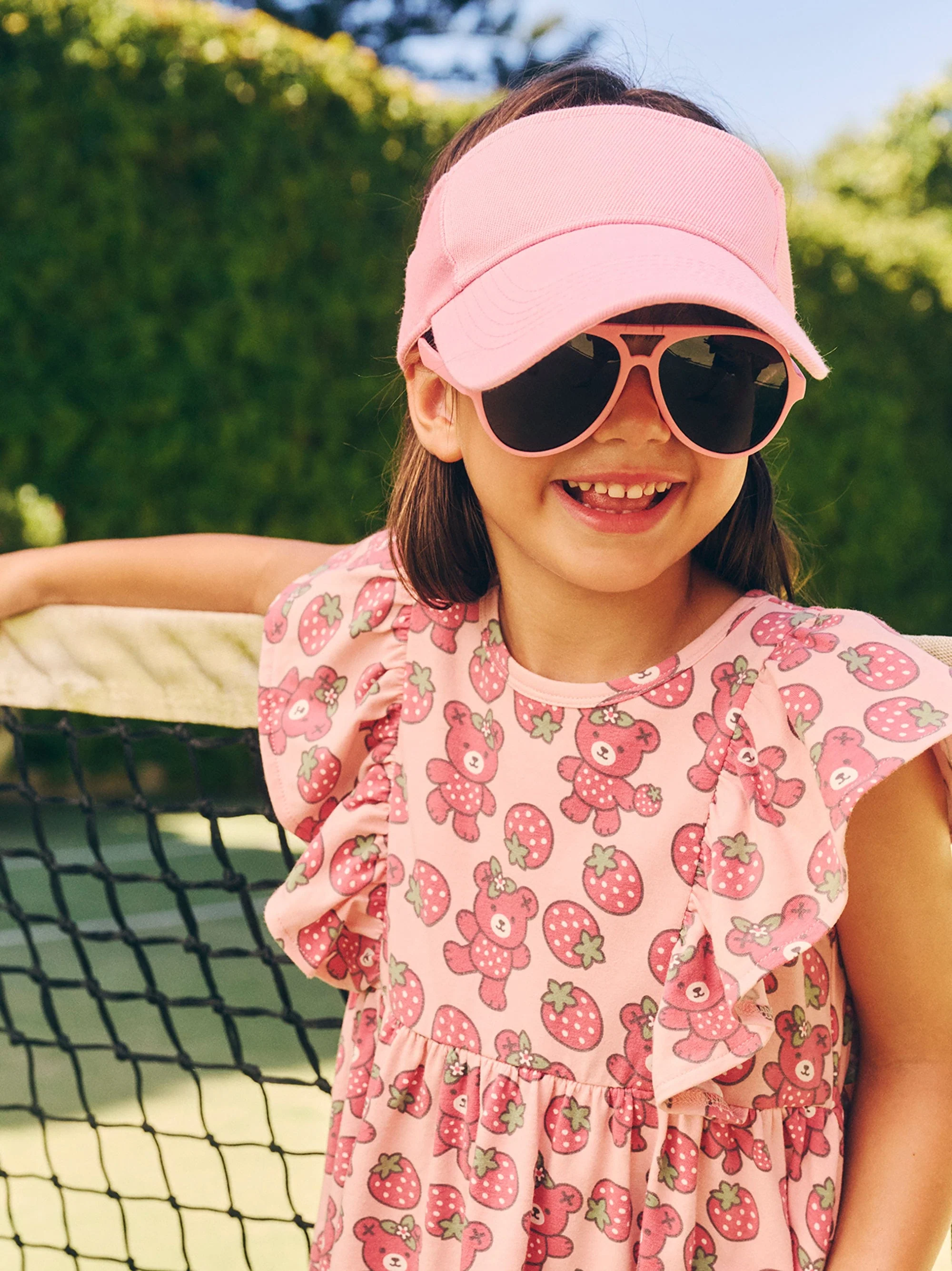 Smiling young girl in pink bear-print dress, visor, and sunglasses stands outdoors near a tennis net on a sunny day.