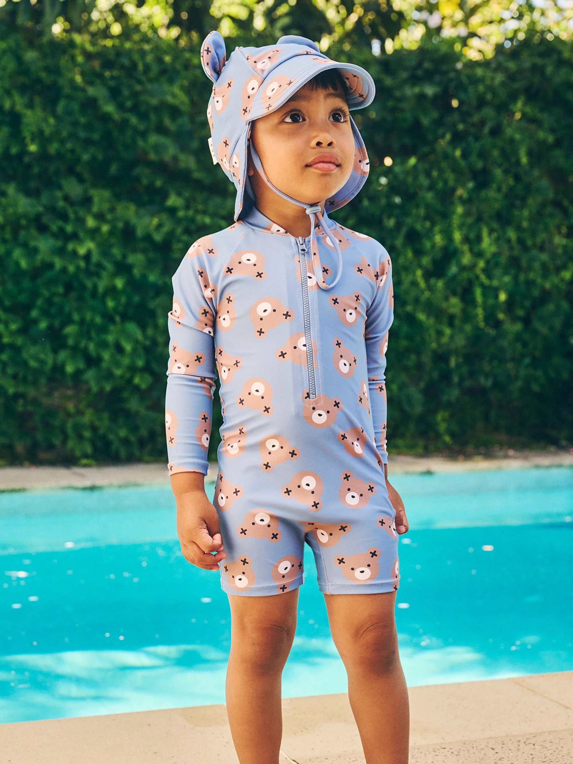 Young child in a blue bear-patterned swimsuit and sun hat stands confidently by a pool, showcasing summer style and outdoor fun.