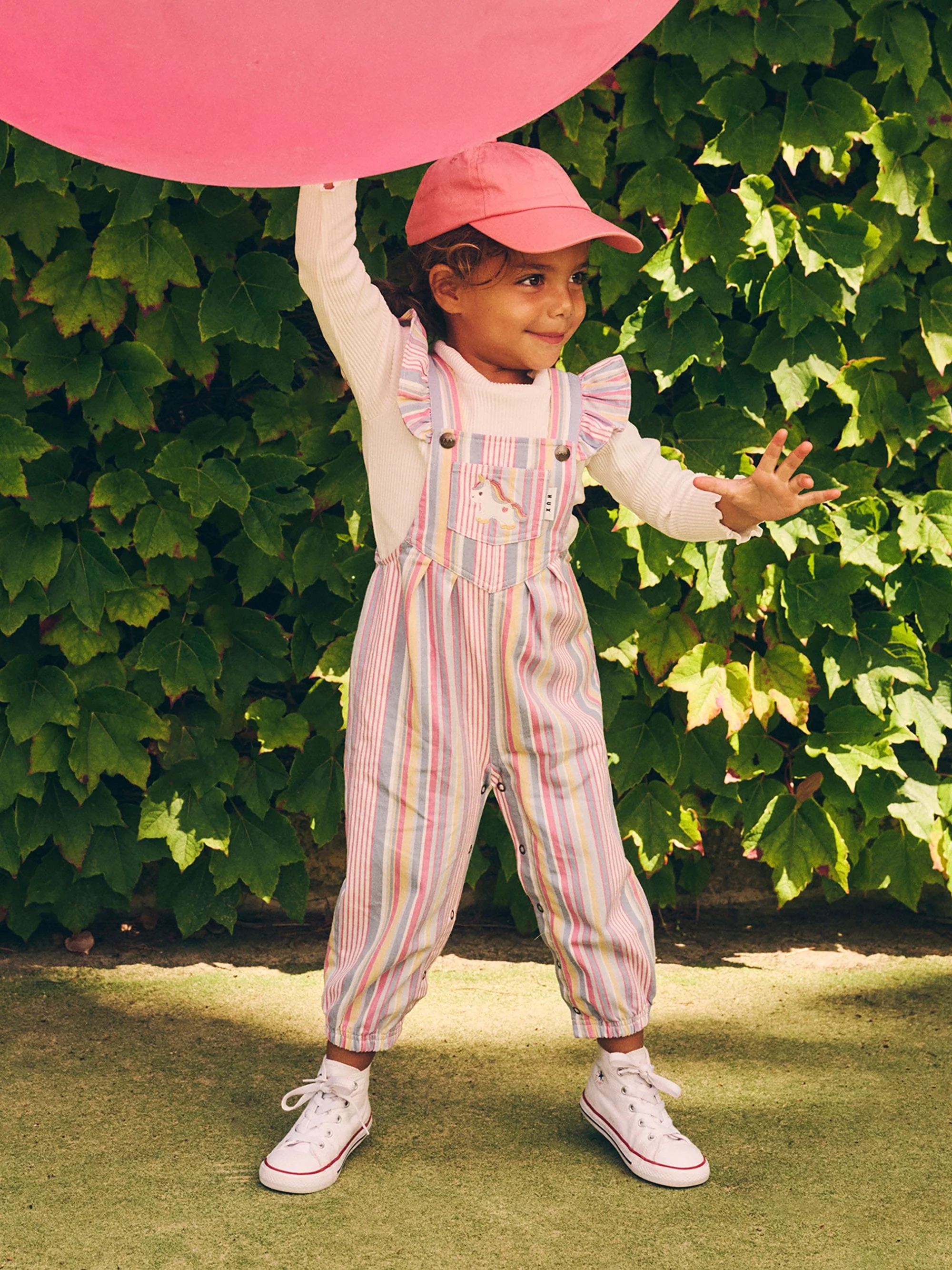 Young child in striped overalls and pink cap playfully reaches for a large pink balloon outdoors against a backdrop of green foliage.