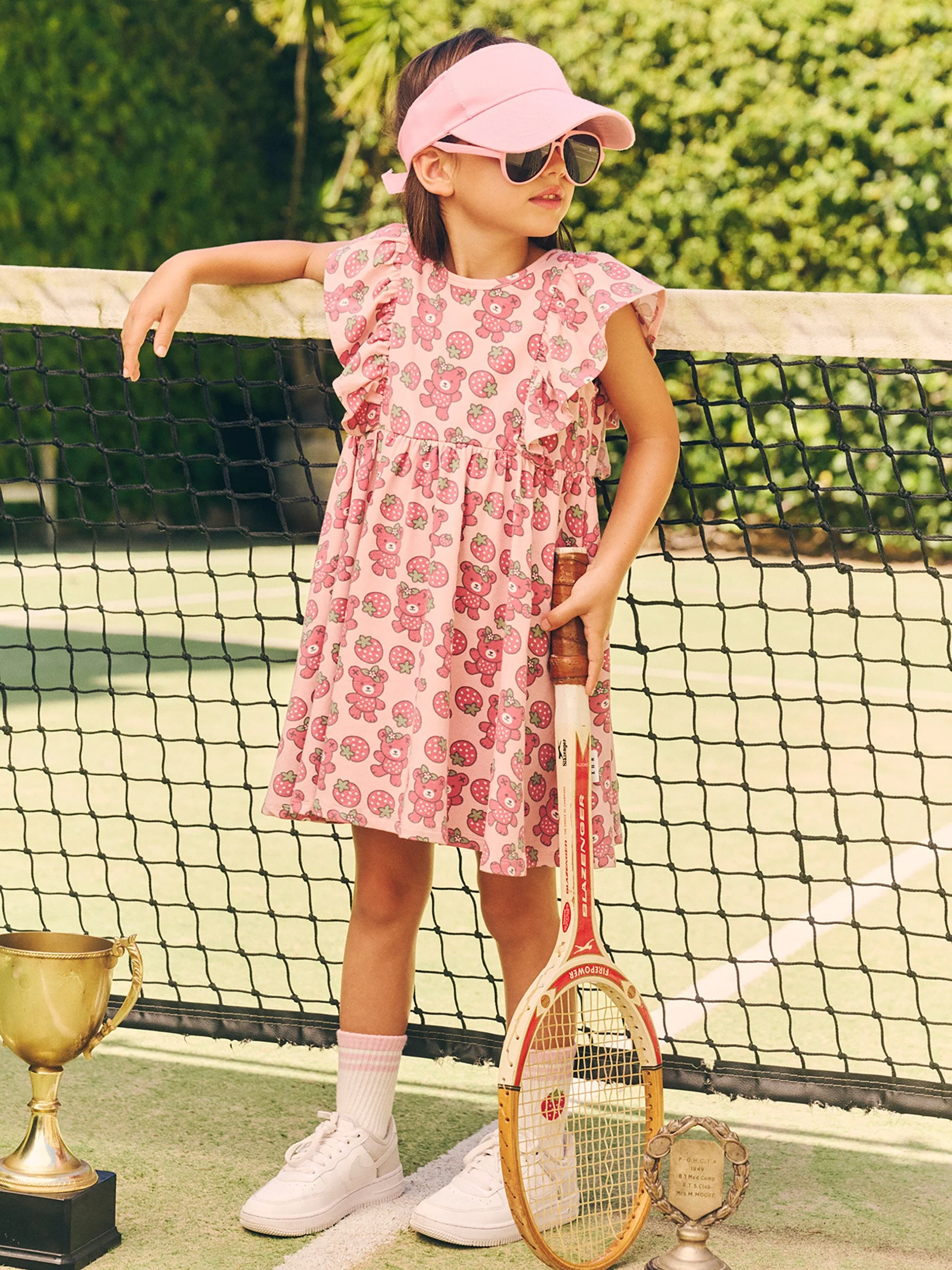 A young girl in a pink dress patterned with teddy bears stands on a tennis court holding a racquet next to two trophies.