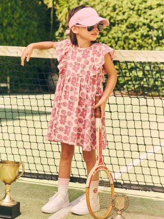 A young girl in a pink dress patterned with teddy bears stands on a tennis court holding a racquet next to two trophies.