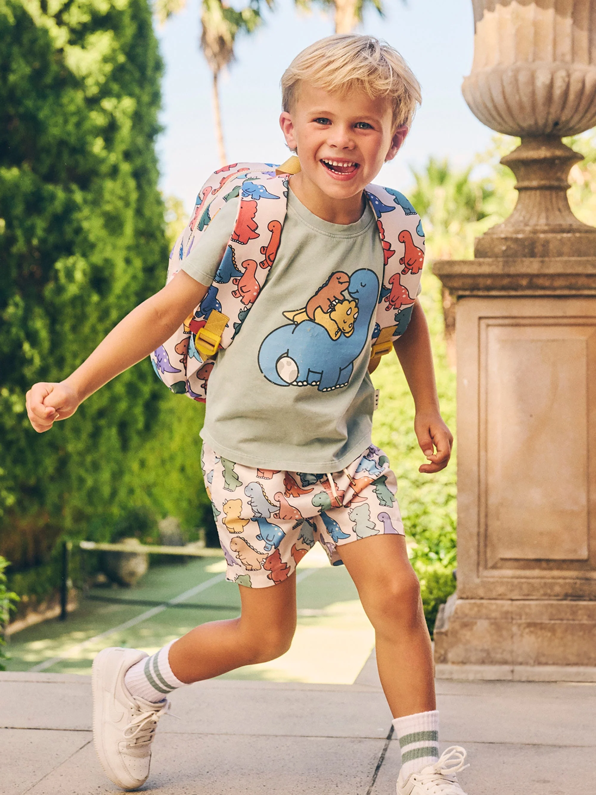 A smiling young boy with blond hair runs outdoors wearing a dinosaur-themed t-shirt, shorts, backpack, and sneakers, on a sunny day.
