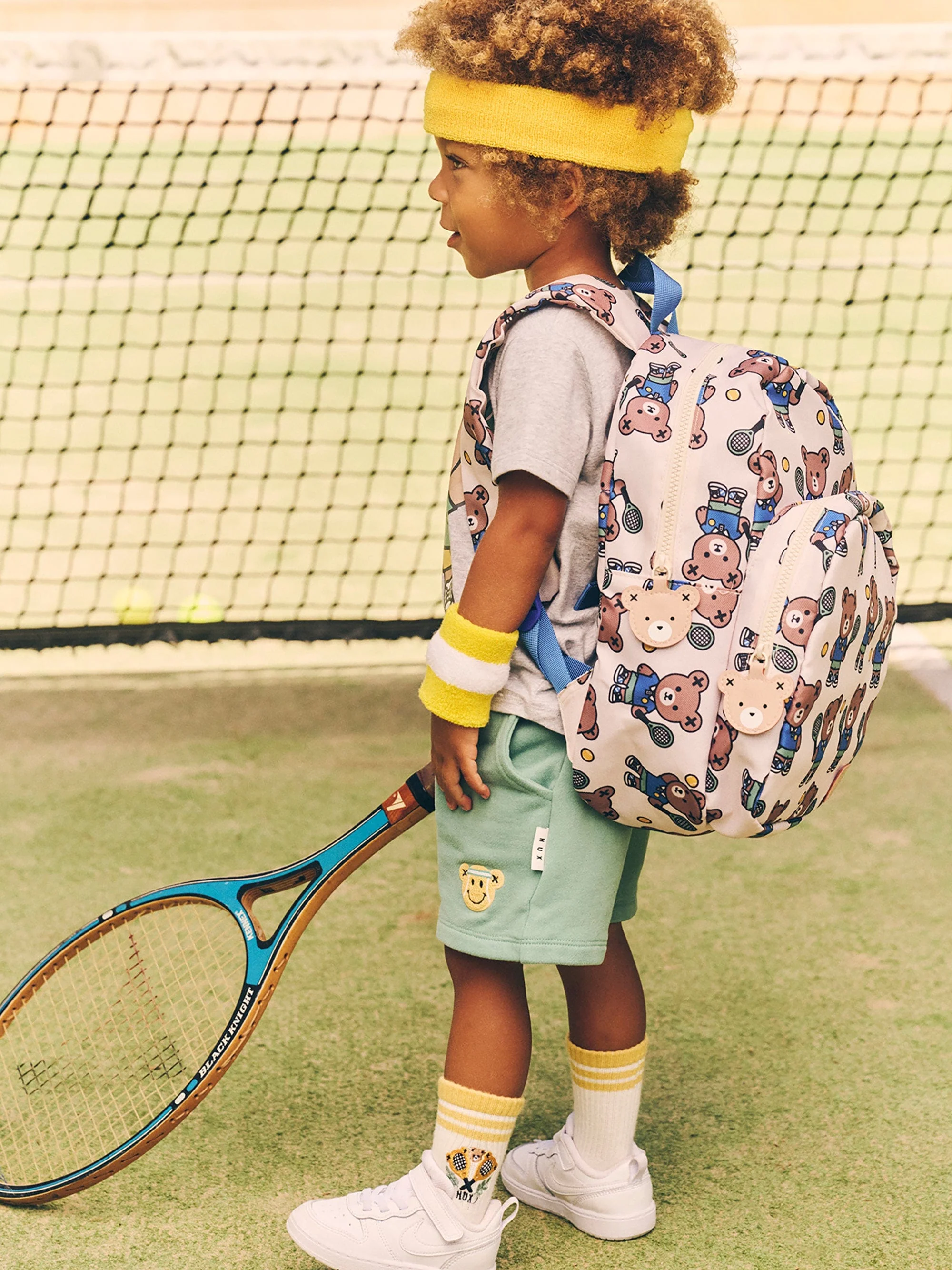 Child wearing a yellow headband and carrying a teddy-bear patterned backpack on a tennis court