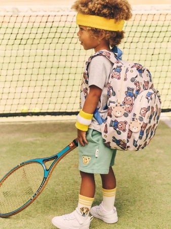 Child wearing a yellow headband and carrying a teddy-bear patterned backpack on a tennis court