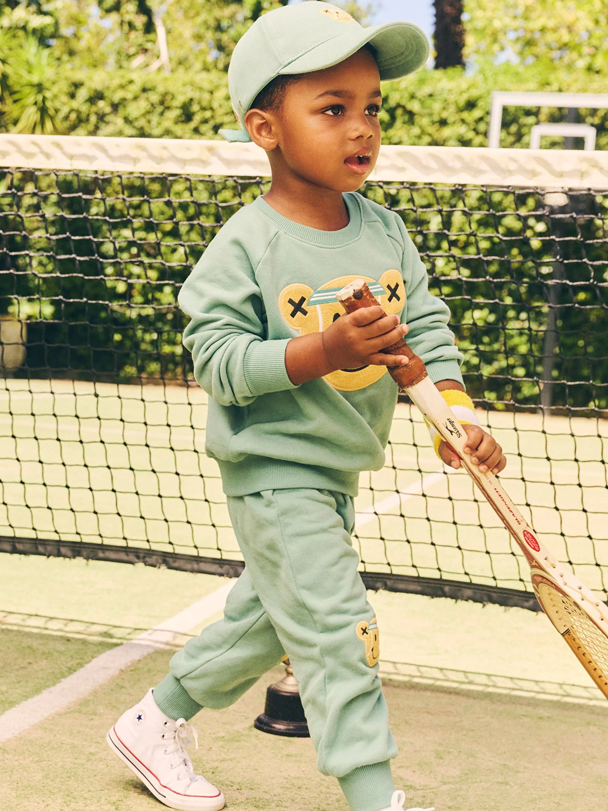 Young child in mint green tracksuit and matching baseball cap holding a tennis racket on a green court with a net backdrop.