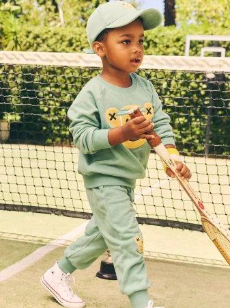 Young child in mint green tracksuit and matching baseball cap holding a tennis racket on a green court with a net backdrop.