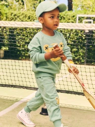 Young child in sage green sweatshirt and matching sweatpants holding a vintage tennis racket near a net