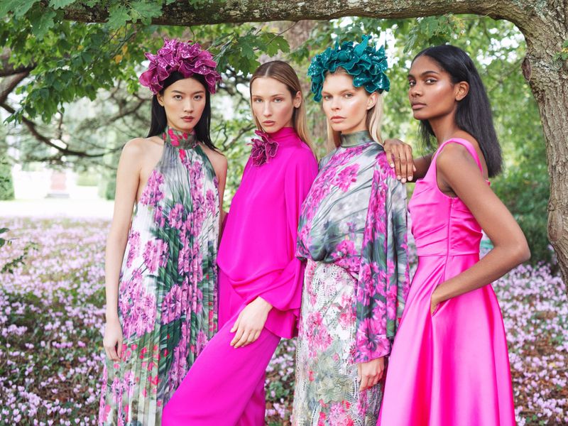 Four female fashion models wearing colorful floral dresses and decorative floral headpieces pose in a garden setting outdoors.