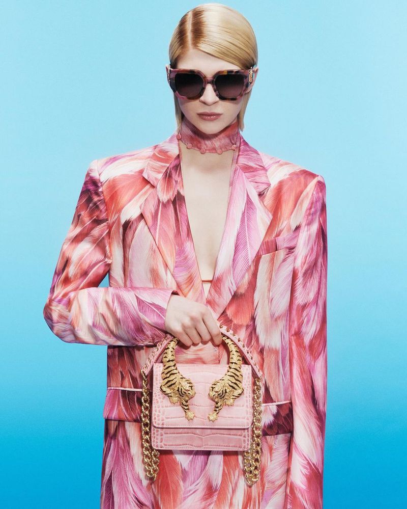 Fashion shot of a woman wearing a pink feather-patterned suit and sunglasses, holding a pink purse with tiger head embellishments against a blue background.