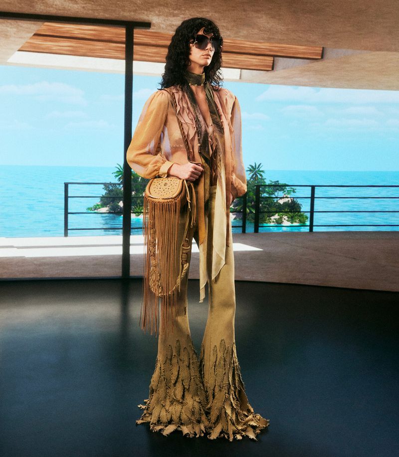 A woman models tan bell-bottom pants with feather details and a matching fringed handbag in front of a blue ocean and island vista.