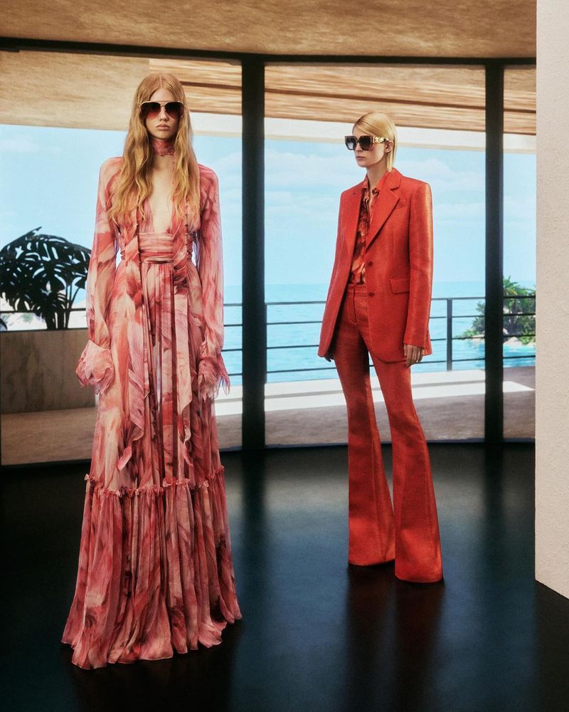 Two women pose in fashionable dresses and sunglasses in a modern setting with large windows and a view of the ocean and greenery.