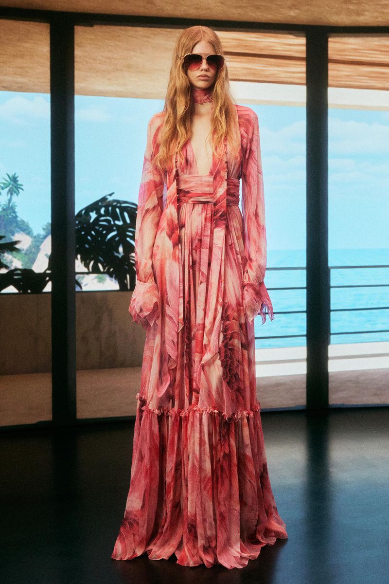 Fashion model in a pink floral chiffon gown with sunglasses poses on a terrace overlooking the ocean in a full-length shot.