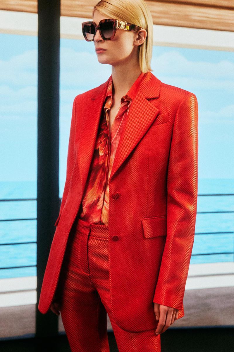 Woman poses in a textured red blazer and pantsuit with a floral shirt. She wears oversized sunglasses against a coastal backdrop.