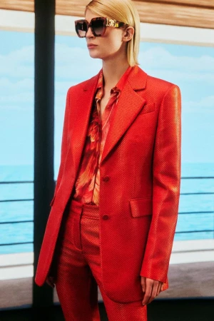 Woman poses in a textured red blazer and pantsuit with a floral shirt. She wears oversized sunglasses against a coastal backdrop.