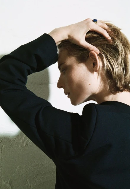 Profile portrait of a fair-skinned adult woman in a dark blazer with her hand on her head in front of a textured white wall.