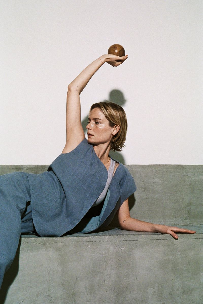 Adult woman in casual blue outfit poses on concrete surface, holding brown sphere overhead against neutral background in a fashion editorial.