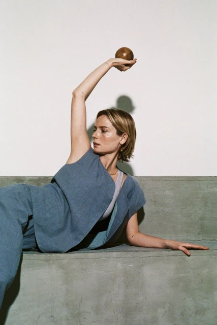 Adult woman in casual blue outfit poses on concrete surface, holding brown sphere overhead against neutral background in a fashion editorial.