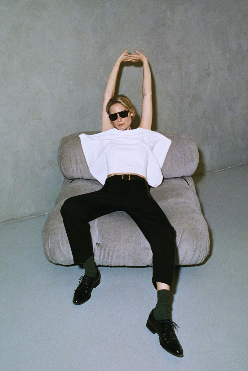 Stylish woman in sunglasses poses on a grey chair against a grey wall, wearing a white shirt and black pants, in a fashion shoot.