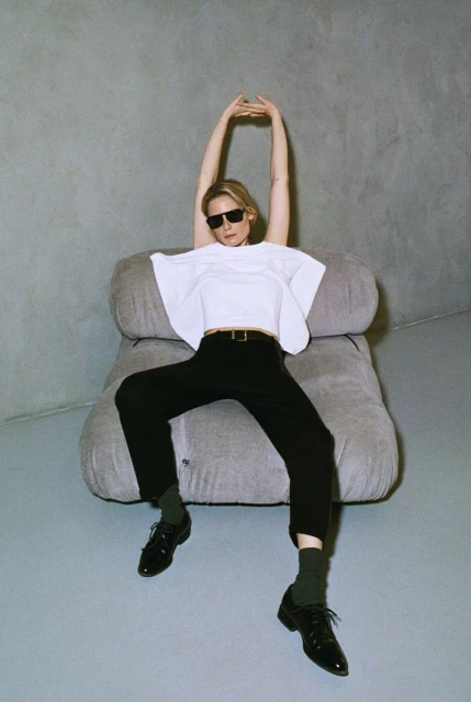 Stylish woman in sunglasses poses on a grey chair against a grey wall, wearing a white shirt and black pants, in a fashion shoot.