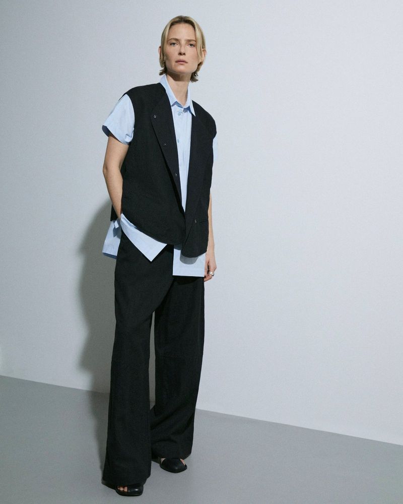 Full-length shot of a blond woman in a black vest and wide-leg pants posing against a neutral background, looking directly at the camera.