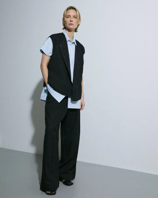 Full-length shot of a blond woman in a black vest and wide-leg pants posing against a neutral background, looking directly at the camera.