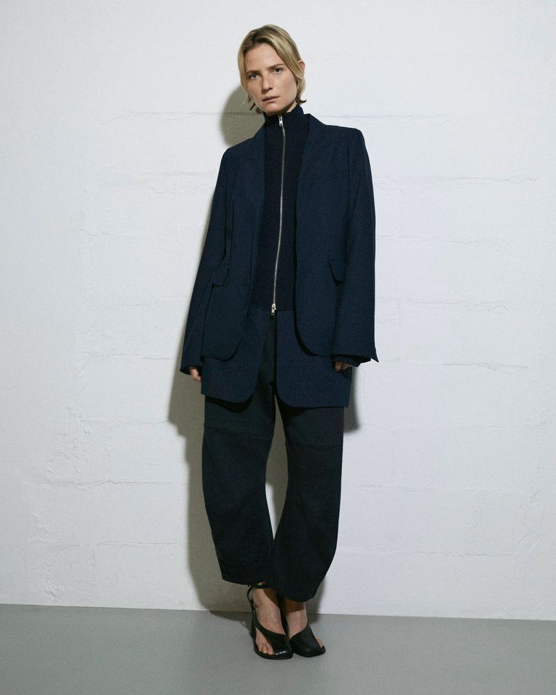 An adult female stands indoors in a dark, oversized blazer and pants, paired with strappy heeled sandals, against a textured white wall.