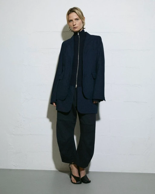 An adult female stands indoors in a dark, oversized blazer and pants, paired with strappy heeled sandals, against a textured white wall.