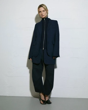 An adult female stands indoors in a dark, oversized blazer and pants, paired with strappy heeled sandals, against a textured white wall.