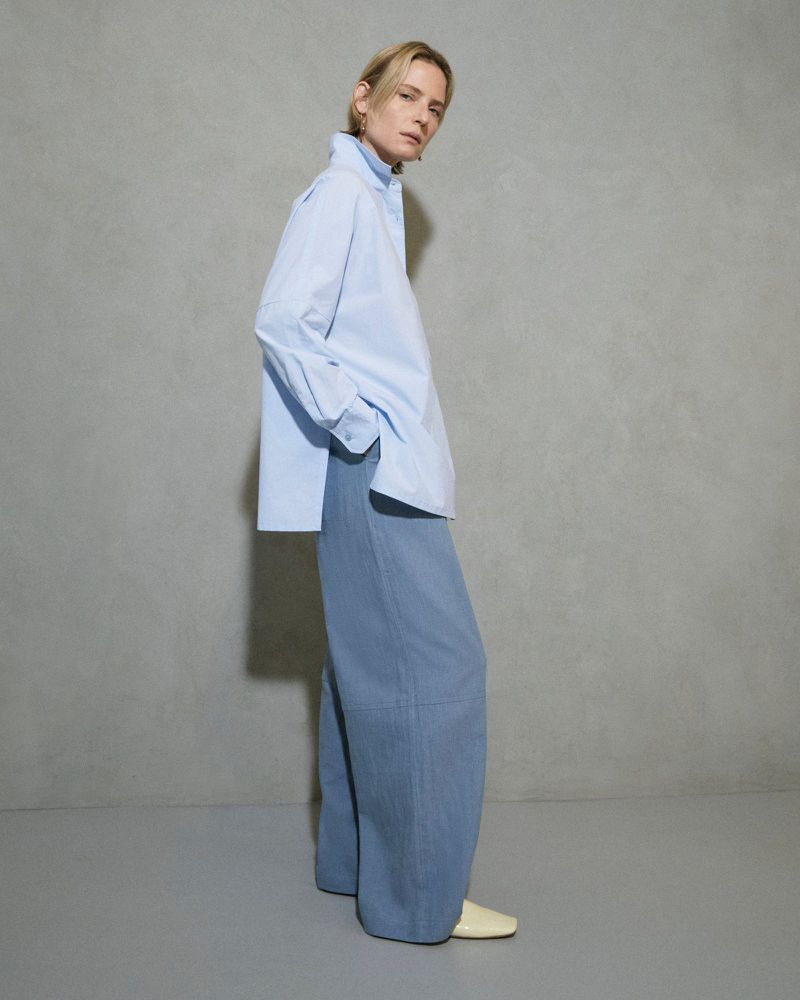 Woman in a blue oversized shirt and wide-legged pants stands in a photo studio with a light grey backdrop for a fashion editorial shoot.