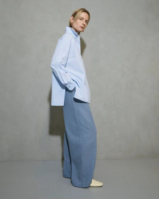 Woman in a blue oversized shirt and wide-legged pants stands in a photo studio with a light grey backdrop for a fashion editorial shoot.
