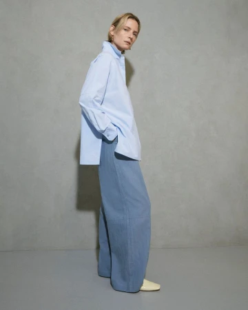 Woman in a blue oversized shirt and wide-legged pants stands in a photo studio with a light grey backdrop for a fashion editorial shoot.