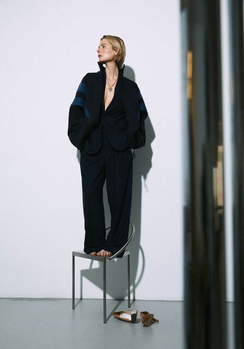 An adult female stands on a metal stool in a fashion pose, wearing a dark blue suit in front of a white wall with nearby shoes.