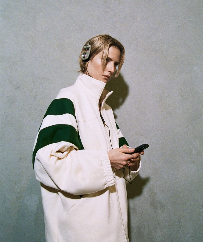 Portrait of a blonde adult woman wearing headphones and an oversized jacket, looking at a black mobile phone in her hands.