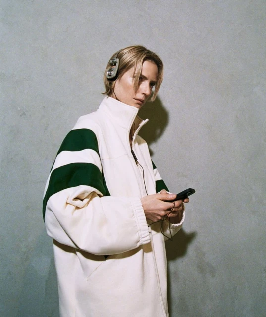 Portrait of a blonde adult woman wearing headphones and an oversized jacket, looking at a black mobile phone in her hands.