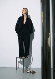 Model standing barefoot on a metal chair wearing a dark suit jacket and trousers with blue sleeve stripes against a white studio wall.
