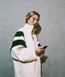 Model wearing white and green track jacket holding a smartphone against a textured concrete wall