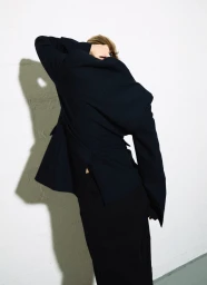 Model with blonde hair wearing a dark blazer over black trousers against a plain white wall with a sharp shadow cast