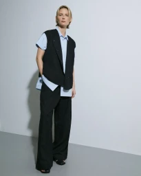 Model standing against a white studio wall wearing a light blue button-down shirt under a dark sleeveless vest with matching trousers.